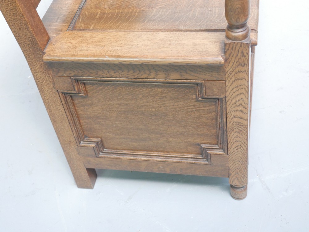 A medium brown oak hall bench featuring a tri-panel backrest, turned armrest supports, and a hinged lid chest base. View XV