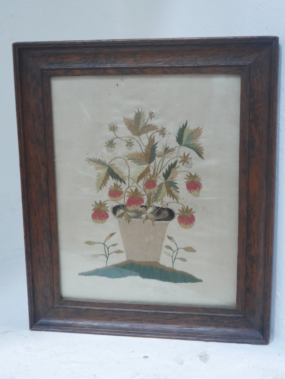 A framed antique needlework picture depicting a potted strawberry plant with red fruit and green leaves on a beige fabric background inside a dark wooden frame.