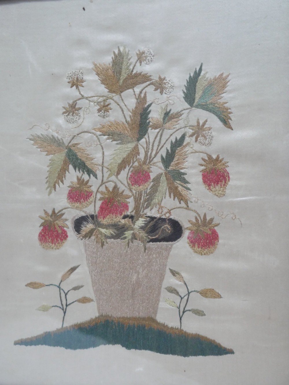 A framed antique needlework picture depicting a potted strawberry plant with red fruit and green leaves on a beige fabric background inside a dark wooden frame. View II