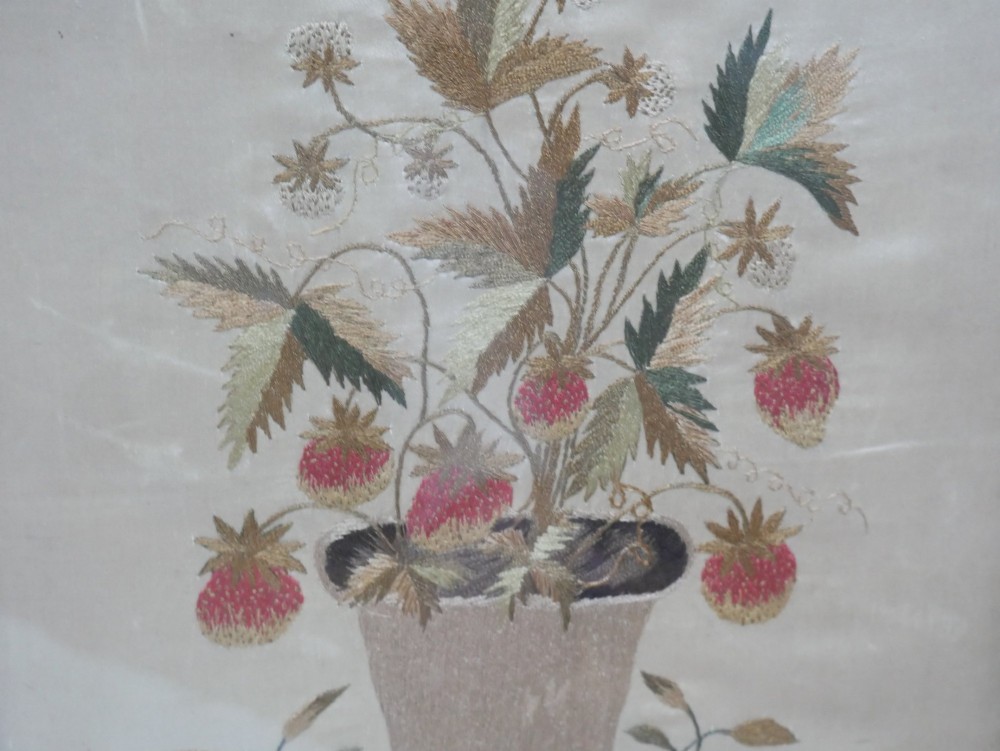 A framed antique needlework picture depicting a potted strawberry plant with red fruit and green leaves on a beige fabric background inside a dark wooden frame. View III