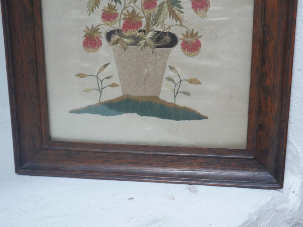A framed antique needlework picture depicting a potted strawberry plant with red fruit and green leaves on a beige fabric background inside a dark wooden frame. View IV