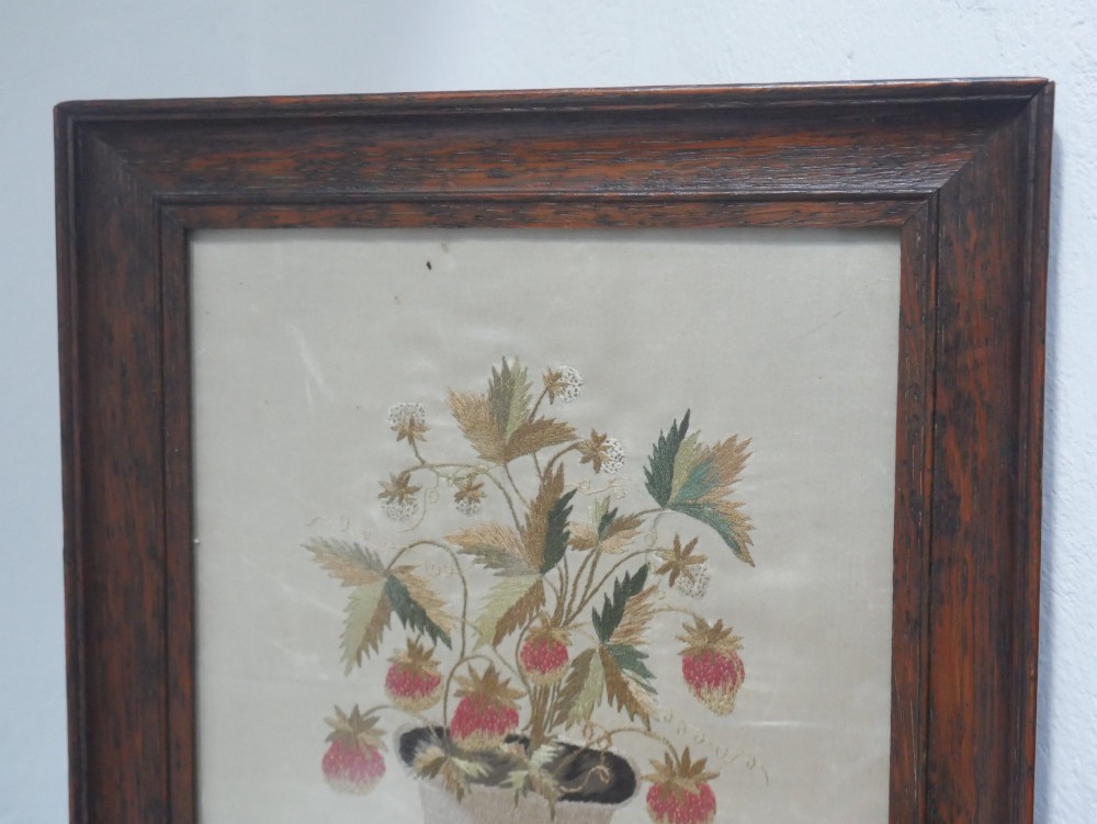 A framed antique needlework picture depicting a potted strawberry plant with red fruit and green leaves on a beige fabric background inside a dark wooden frame. View V