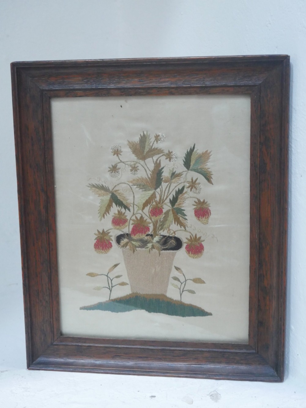 A framed antique needlework picture depicting a potted strawberry plant with red fruit and green leaves on a beige fabric background inside a dark wooden frame. View VI