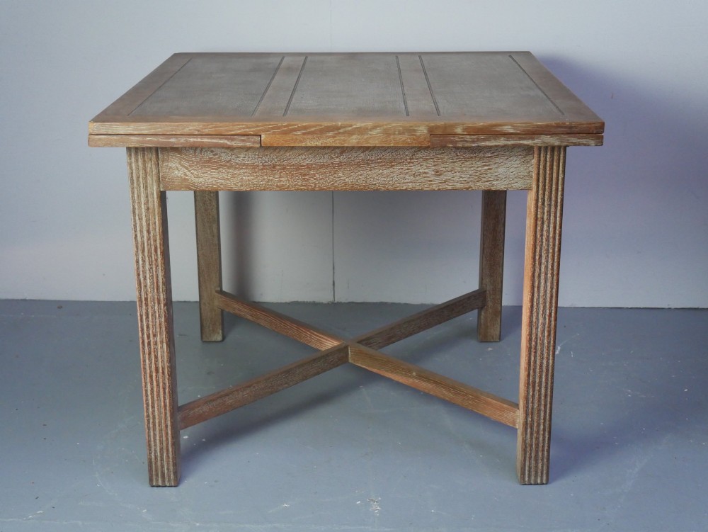 A square limed oak dining table featuring reeded legs, an X-shaped cross stretcher, and a draw-leaf top mechanism.