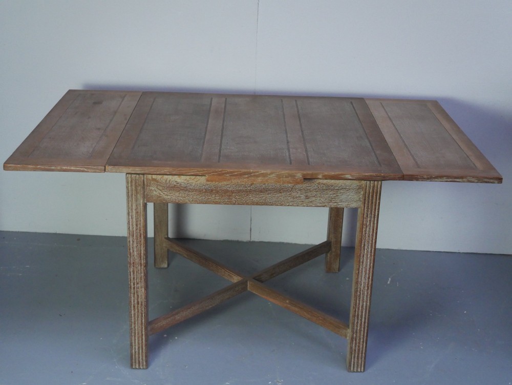 A square limed oak dining table featuring reeded legs, an X-shaped cross stretcher, and a draw-leaf top mechanism. View VII