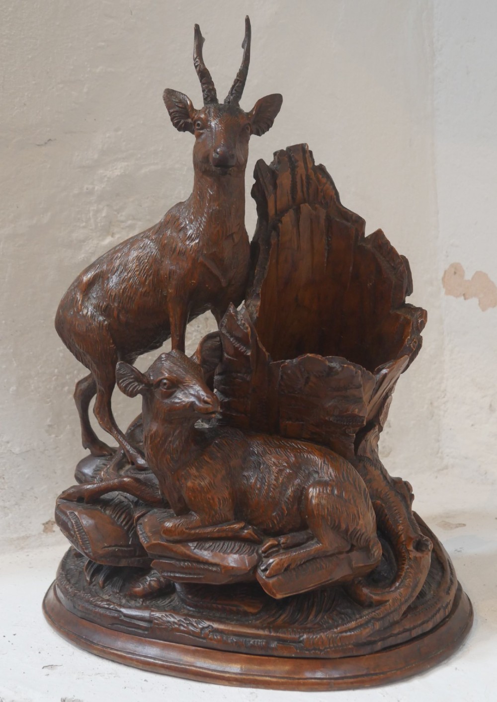 A hand-carved dark wood Black Forest sculpture depicting two mountain goats positioned beside a hollow tree stump on an oval base.
