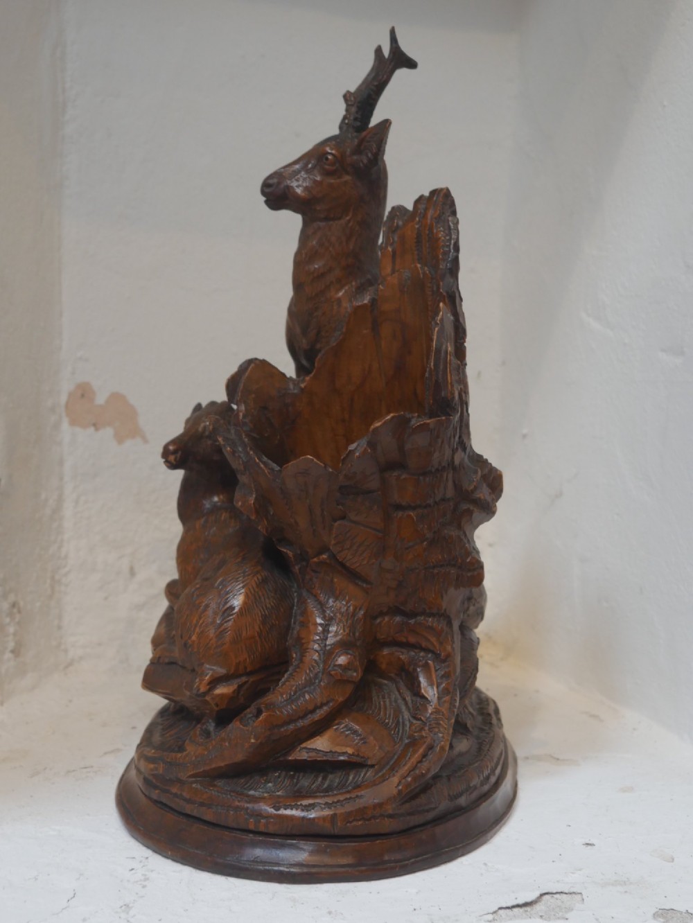 A hand-carved dark wood Black Forest sculpture depicting two mountain goats positioned beside a hollow tree stump on an oval base. View VIII