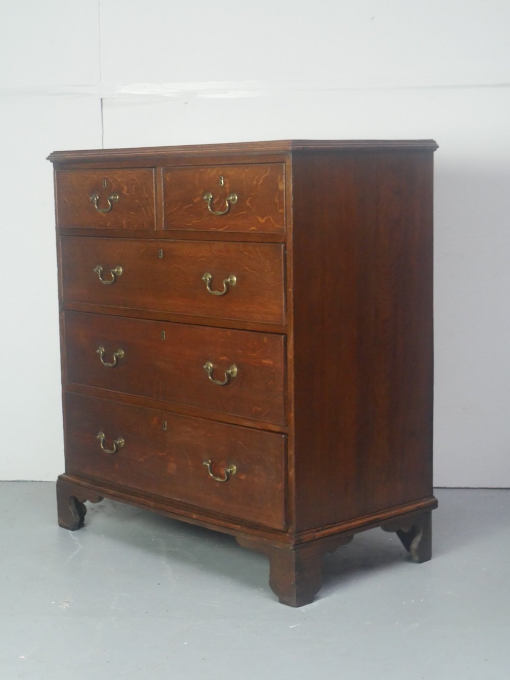 A Georgian oak chest of drawers featuring two short drawers over three long graduated drawers with brass swan-neck handles and bracket feet. View XI
