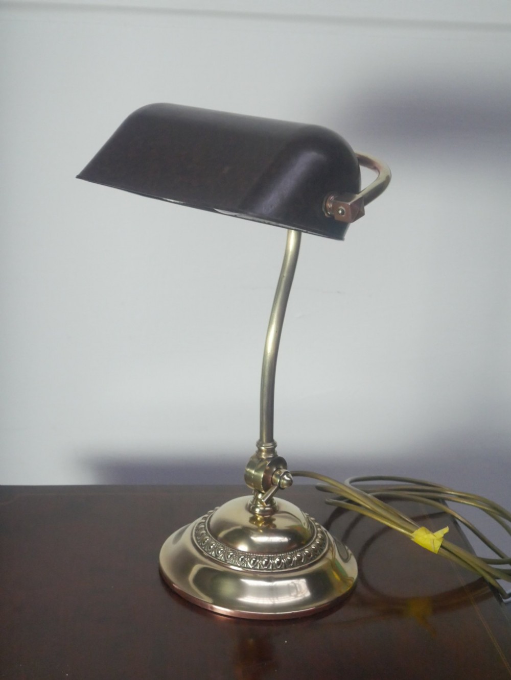 A vintage brass bankers desk lamp featuring a weighted round base, curved stem, and adjustable dark metal shade.