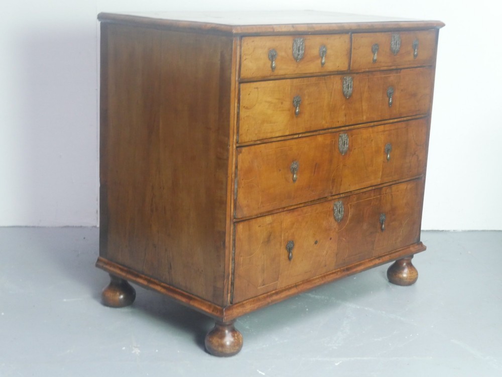 An early 18th century walnut chest of drawers featuring geometric line inlay, brass drop handles, decorative escutcheons, and turned bun feet.