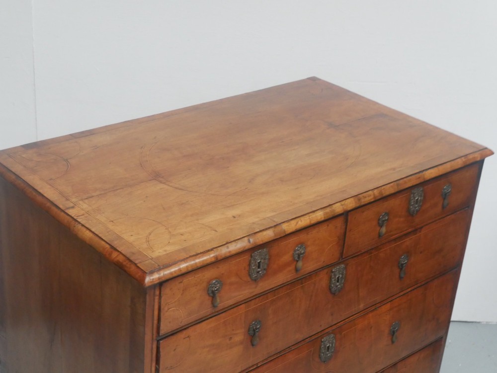 An early 18th century walnut chest of drawers featuring geometric line inlay, brass drop handles, decorative escutcheons, and turned bun feet. View V