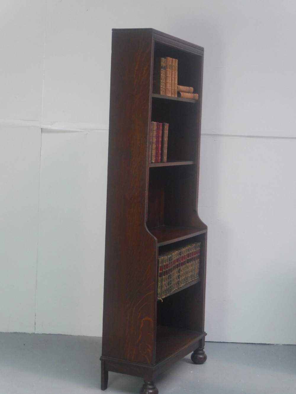 A tall, narrow 1920s dark oak open bookcase featuring five shelves and turned bun feet. View VII
