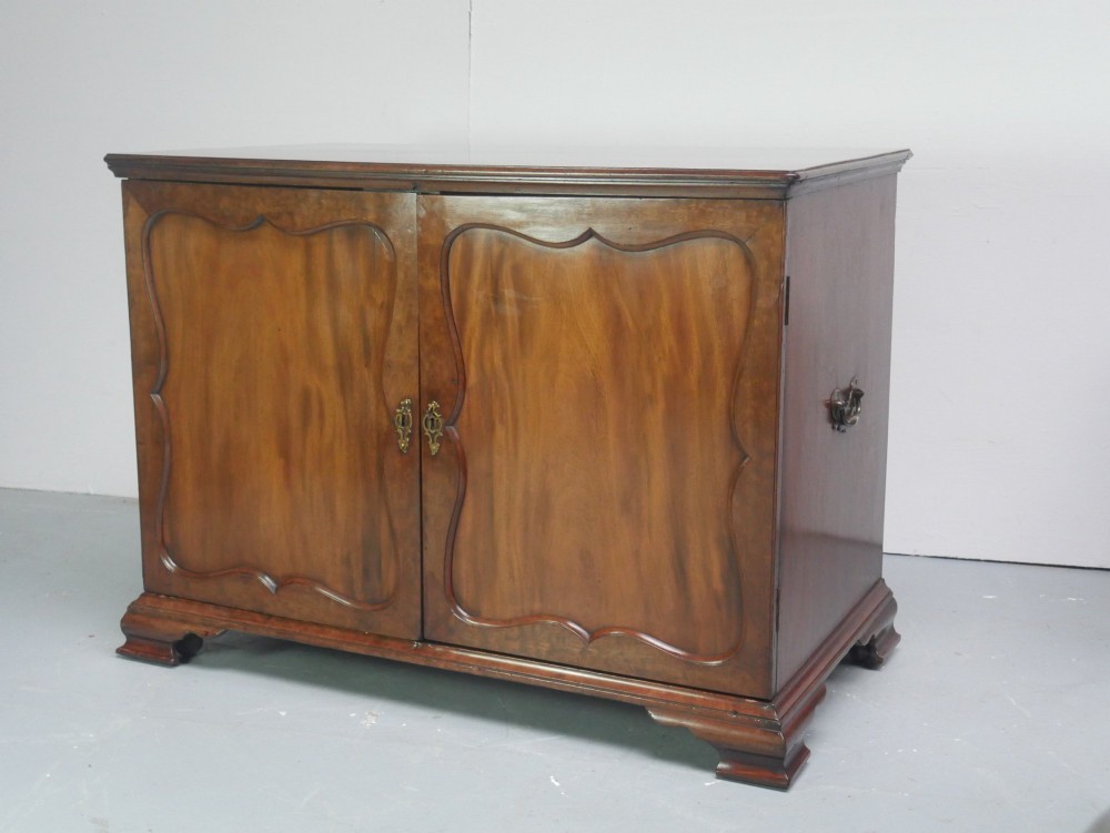 A low Georgian mahogany linen cupboard featuring two shaped panel doors, brass keyhole escutcheons, side handles, and bracket feet.