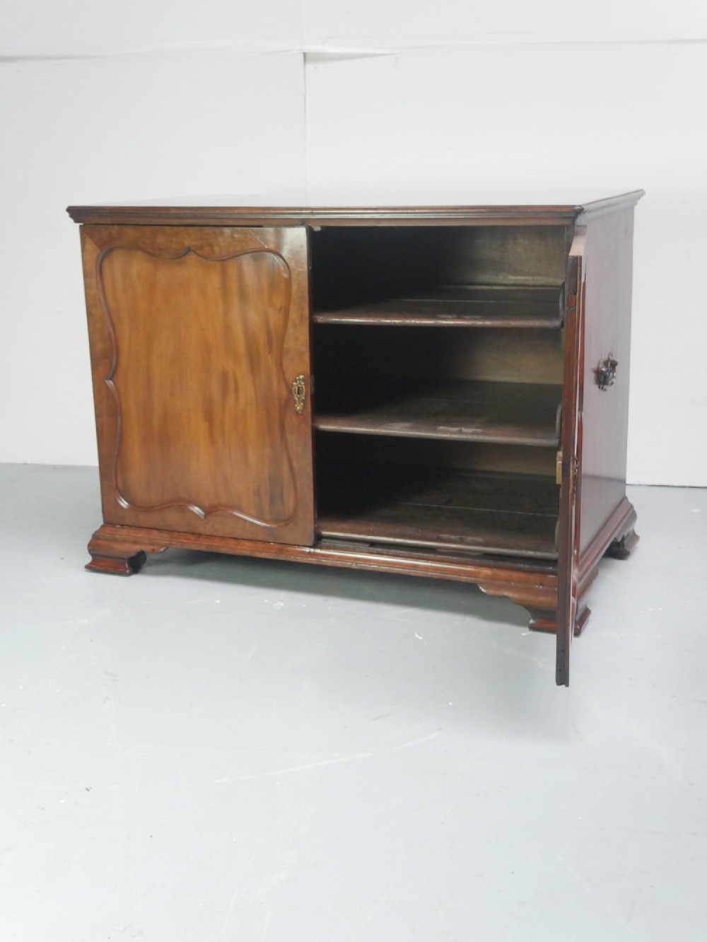 A low Georgian mahogany linen cupboard featuring two shaped panel doors, brass keyhole escutcheons, side handles, and bracket feet. View VIII