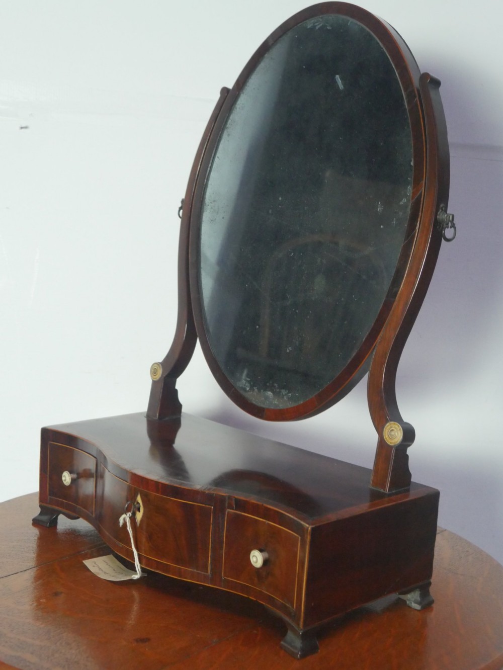 An antique Georgian mahogany dressing mirror with an oval adjustable glass supported on curved arms above a serpentine three-drawer base.