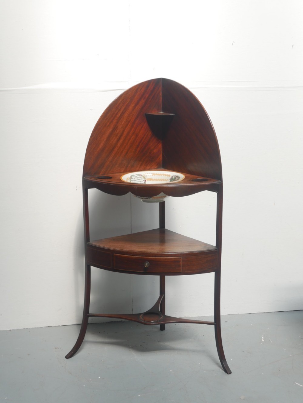 A Georgian mahogany corner washstand featuring a high curved splashback, circular cutouts with a fitted ceramic basin, a middle drawer tier, and splayed legs.
