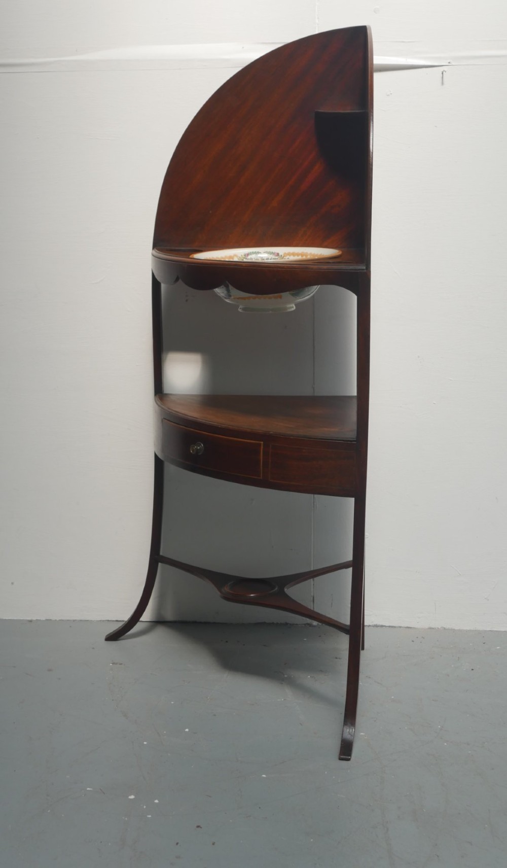 A Georgian mahogany corner washstand featuring a high curved splashback, circular cutouts with a fitted ceramic basin, a middle drawer tier, and splayed legs. View VI