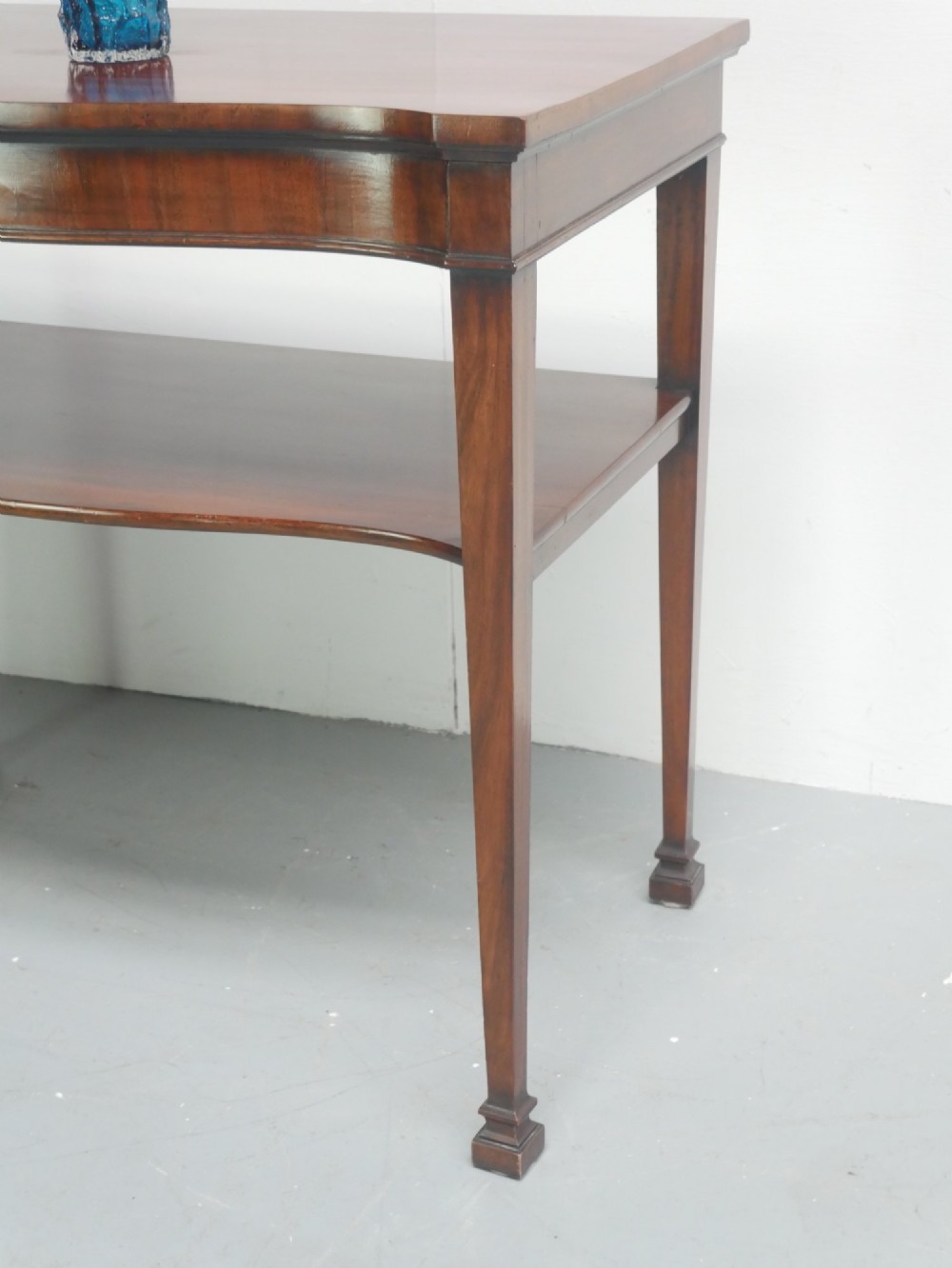 A mahogany console table featuring a shaped top, a lower tier shelf, and tapered square legs. View II