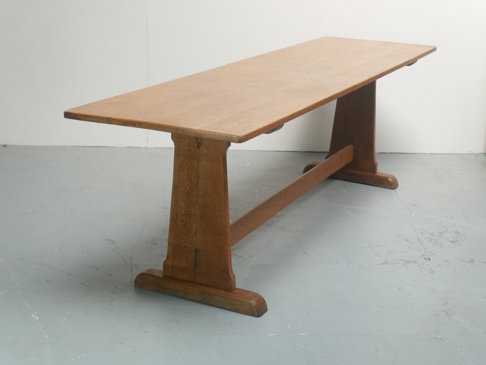 A solid warm brown oak refectory dining table featuring a rectangular plank top, flat trestle leg supports, and a low central stretcher.