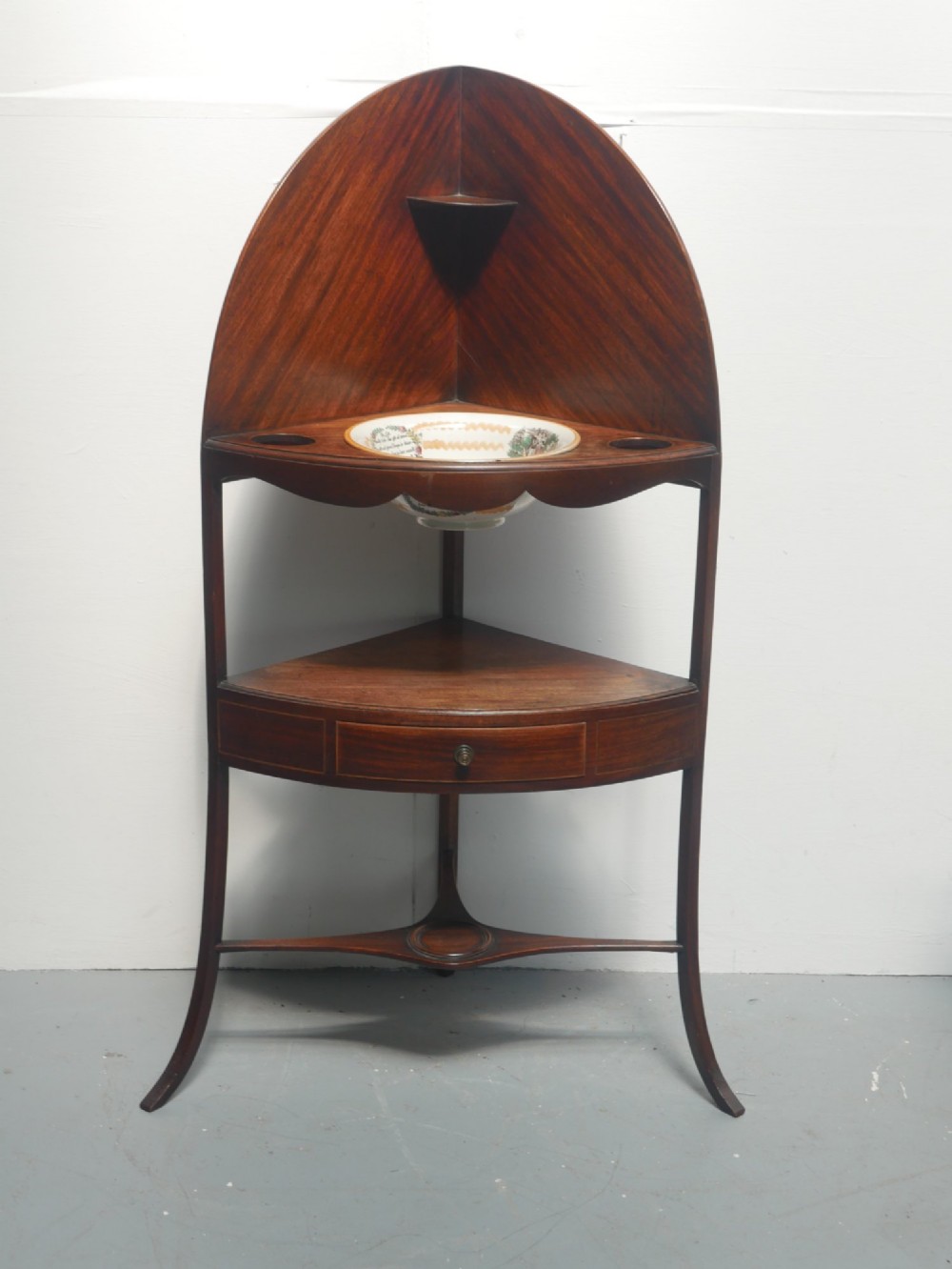 A Georgian mahogany corner washstand featuring a high curved splashback, circular cutouts with a fitted ceramic basin, a middle drawer tier, and splayed legs. View X