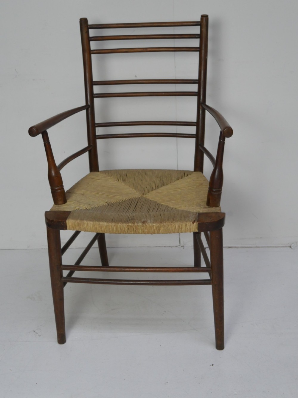 An Arts and Crafts style dark wood armchair featuring a multi-slat ladder back, slender curved armrests, and a hand-woven natural rush seat.