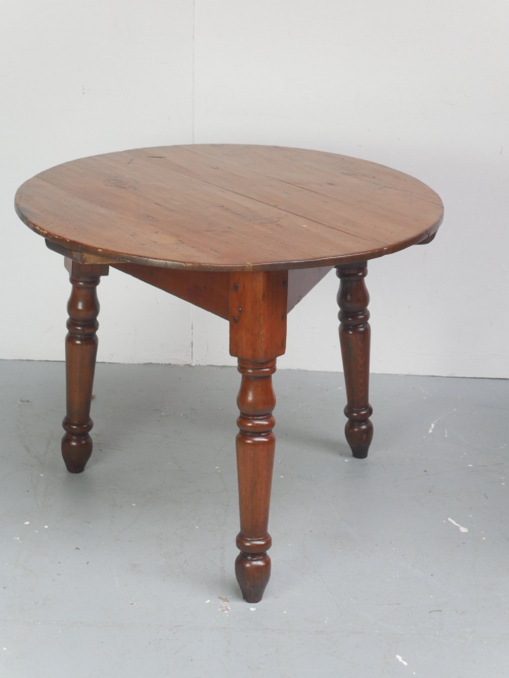 A circular pine cricket table featuring three turned legs joined by a deep triangular apron in a warm brown timber finish. View IX