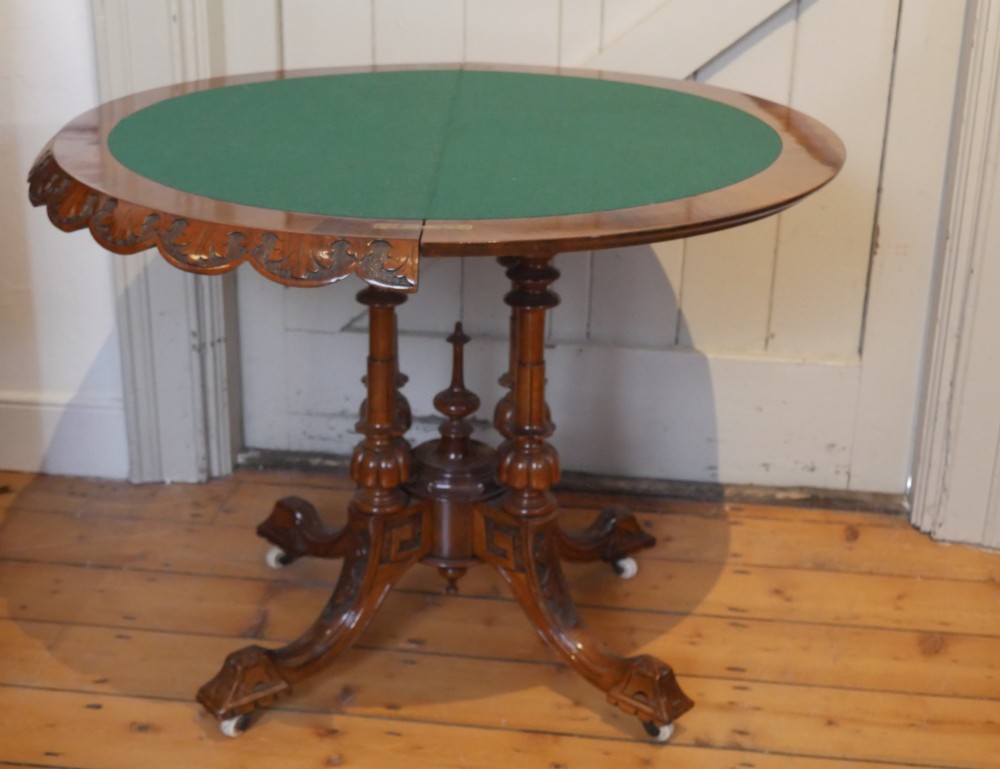 A 19th-century Irish walnut fold-over games table with a scalloped carved apron, turned twin pedestal supports, and carved splayed legs on white castors. View VI