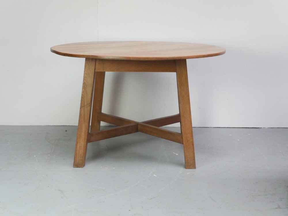 A circular solid oak centre table featuring splayed square legs connected by a central cross-stretcher.