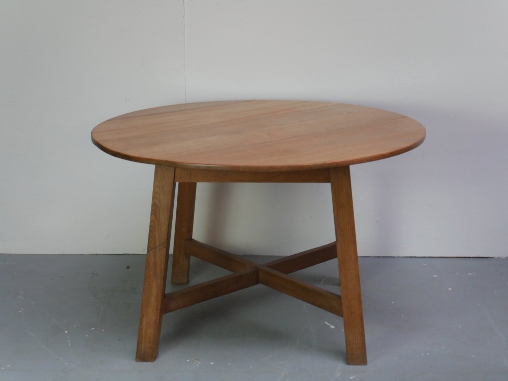 A circular solid oak centre table featuring splayed square legs connected by a central cross-stretcher. View VII