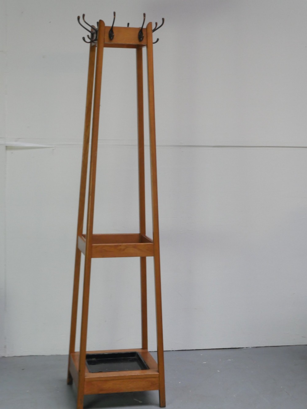 A tall tapered solid oak coat and umbrella stand featuring dark metal hooks around the top and a square metal drip tray set into the wooden base. View VII