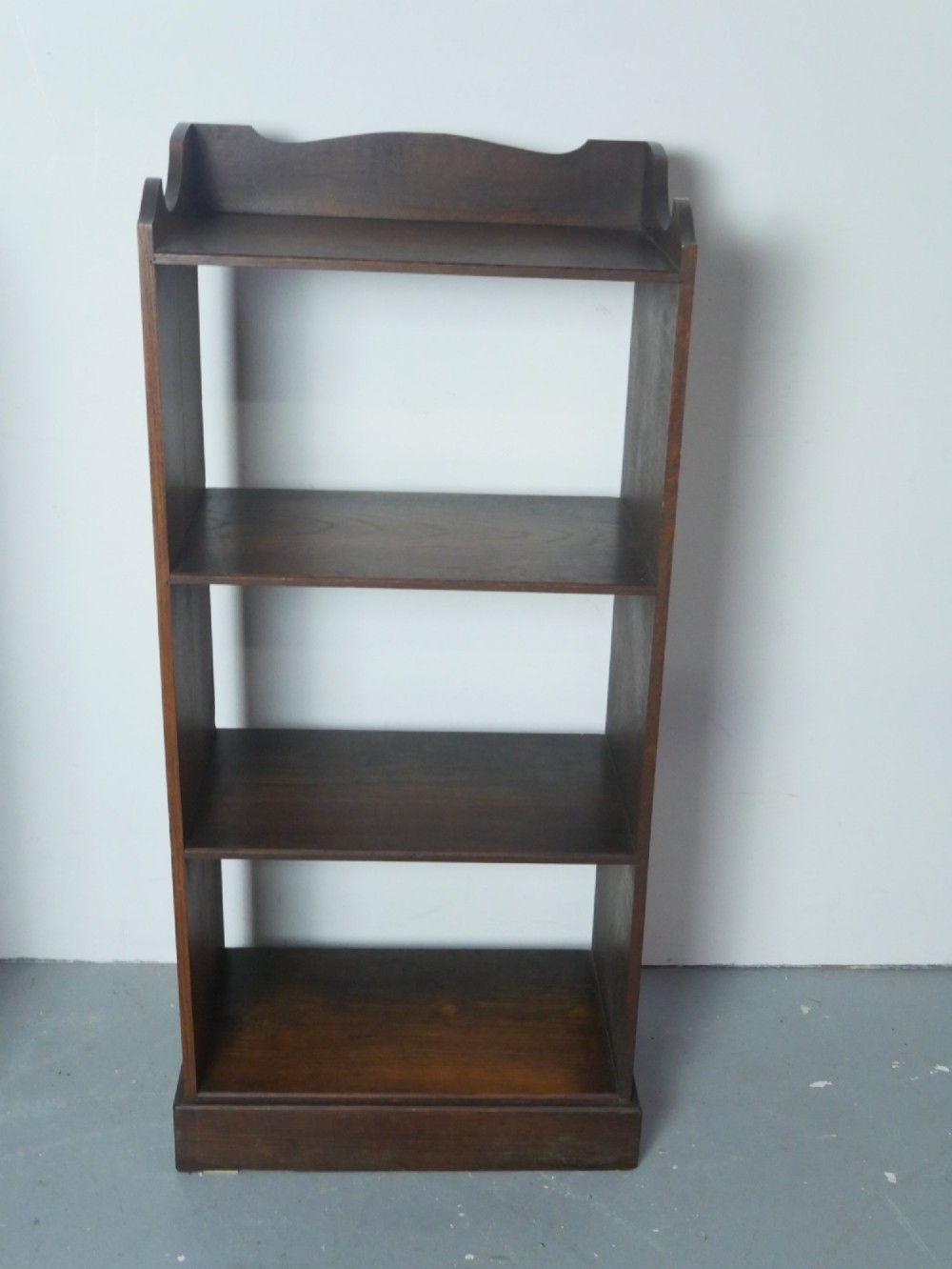 A narrow dark oak open bookcase with four fixed shelves, a shaped gallery top upstand, and a solid plinth base.