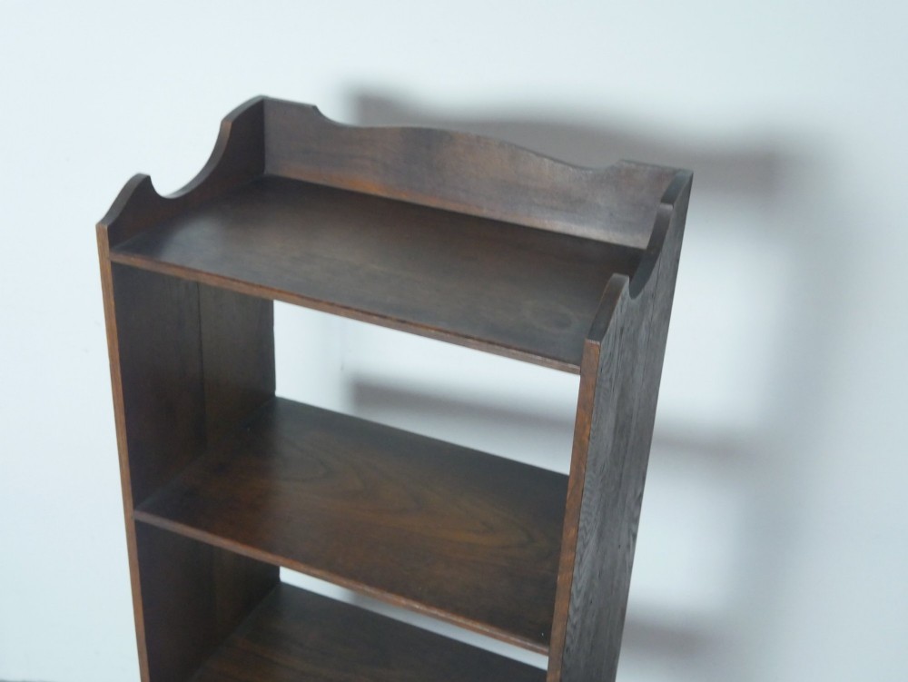 A narrow dark oak open bookcase with four fixed shelves, a shaped gallery top upstand, and a solid plinth base. View III