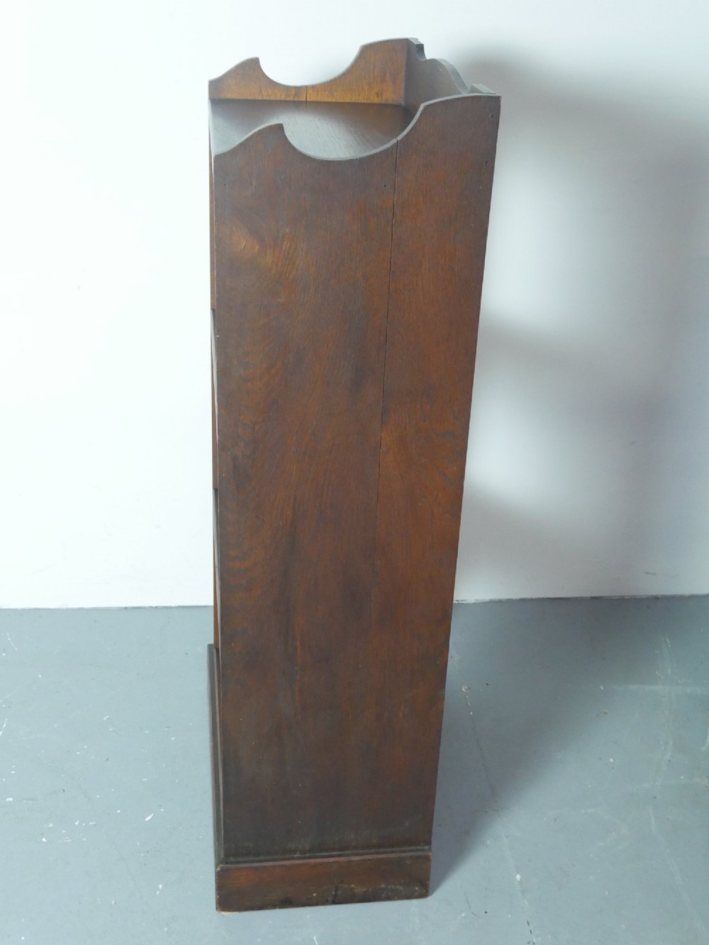 A narrow dark oak open bookcase with four fixed shelves, a shaped gallery top upstand, and a solid plinth base. View V