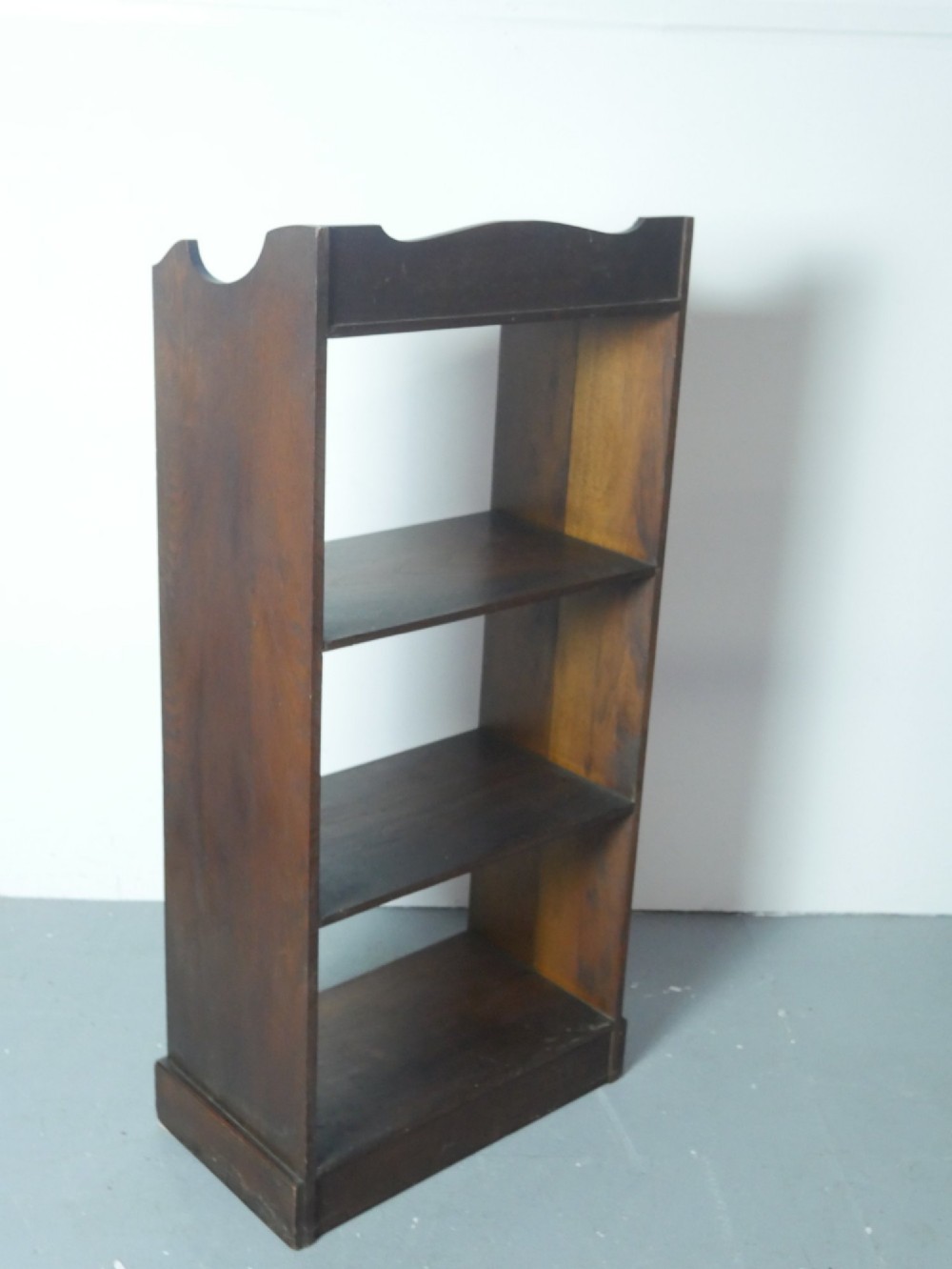 A narrow dark oak open bookcase with four fixed shelves, a shaped gallery top upstand, and a solid plinth base. View VI