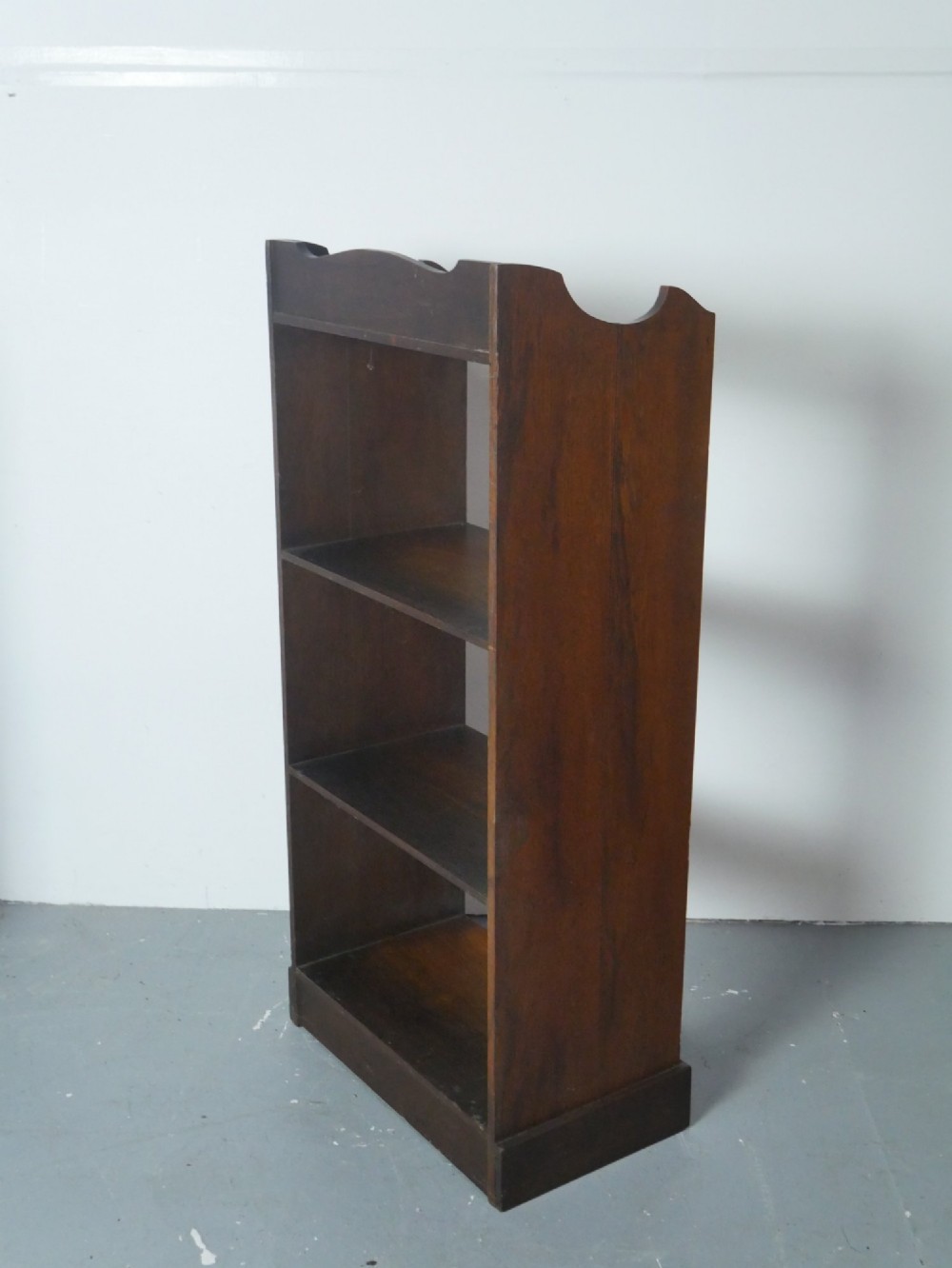 A narrow dark oak open bookcase with four fixed shelves, a shaped gallery top upstand, and a solid plinth base. View VII