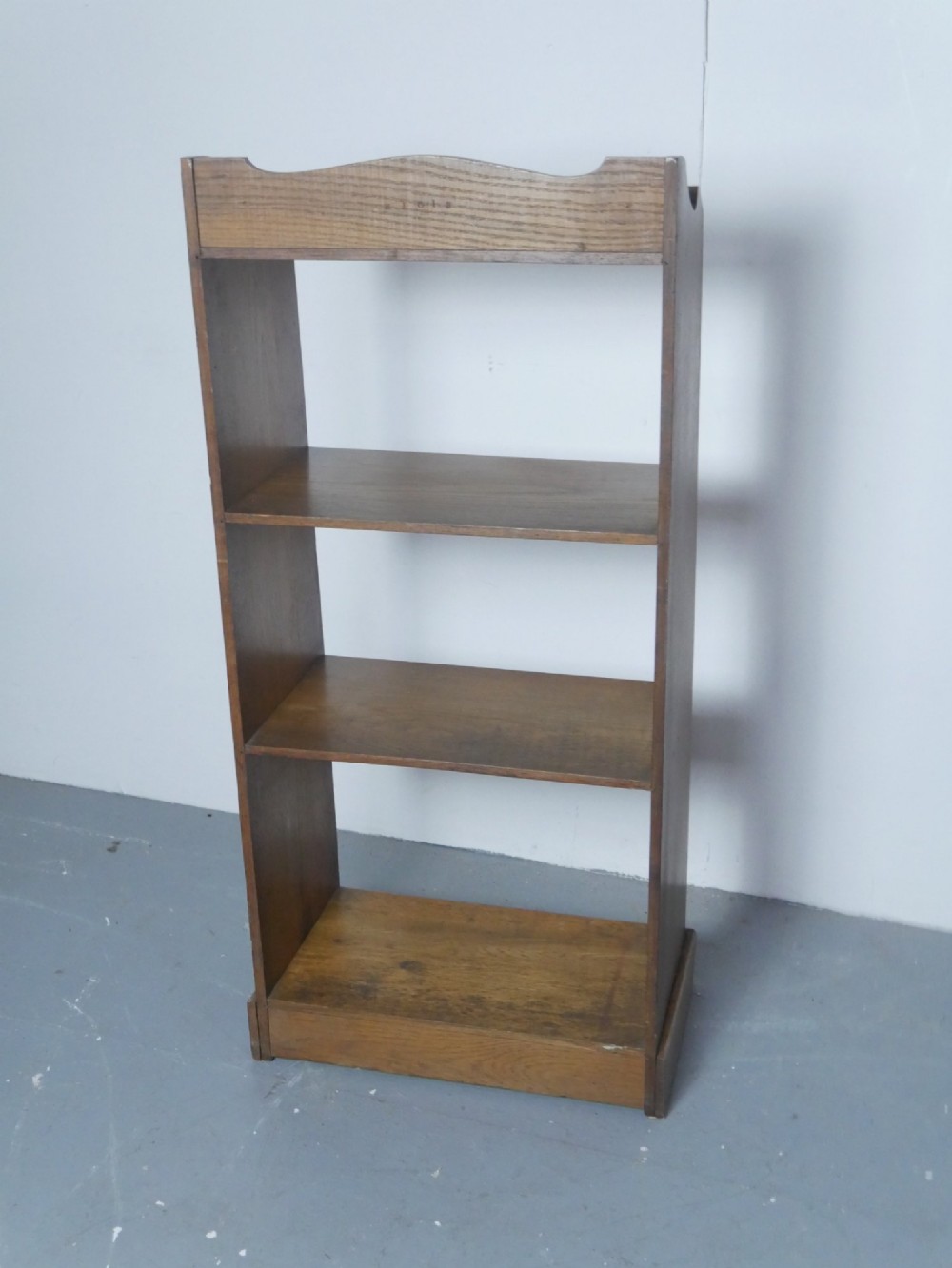 A tall, narrow freestanding open oak bookcase with four shelves, a plinth base, and a shaped top gallery. View V