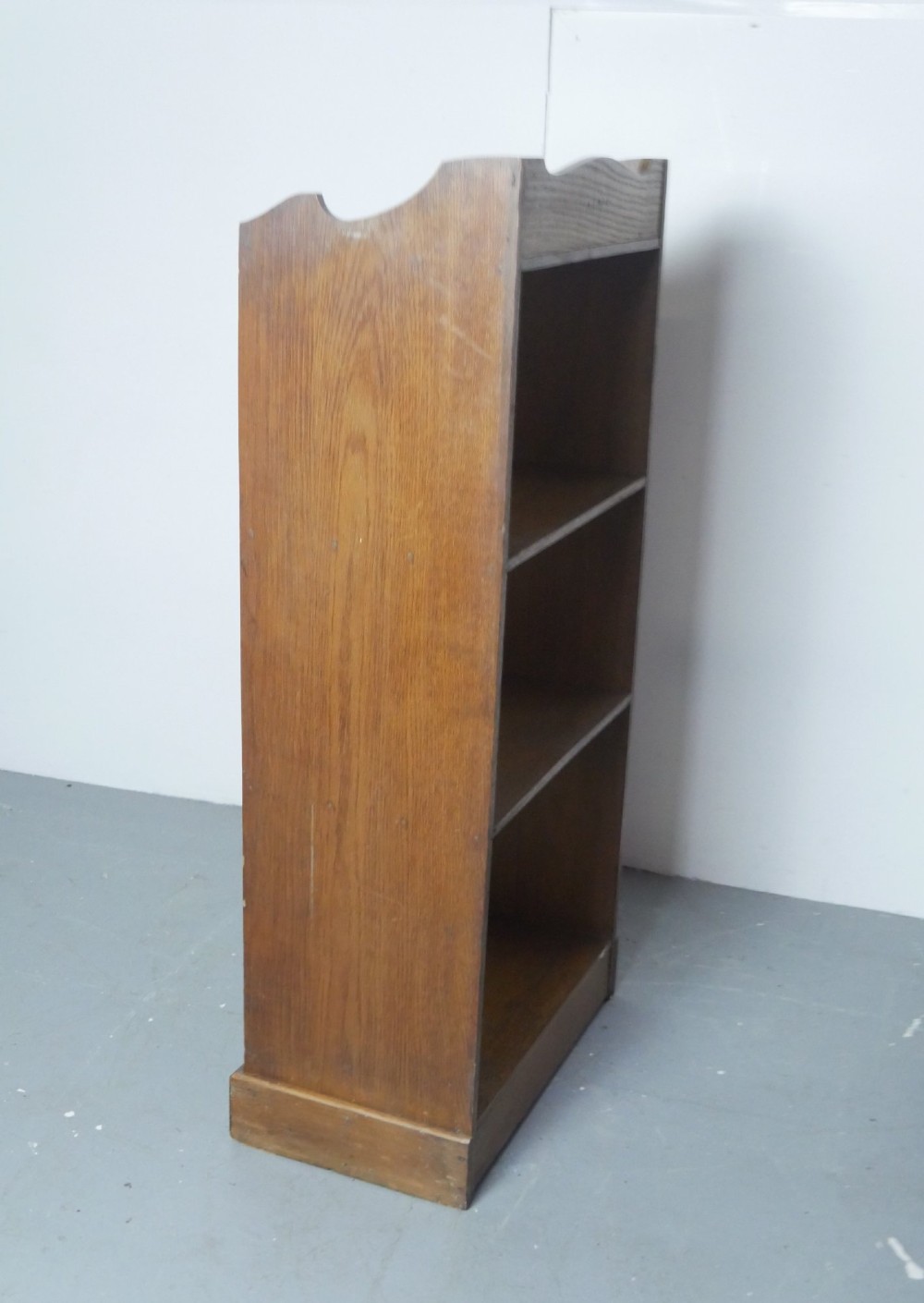 A tall, narrow freestanding open oak bookcase with four shelves, a plinth base, and a shaped top gallery. View VI