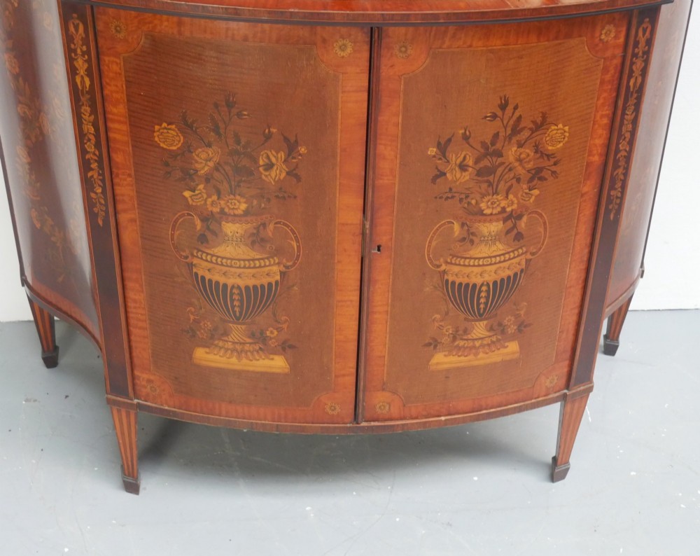 An antique mahogany display cabinet featuring marquetry urn inlays on lower doors, curved bowfront glass doors, glass shelves, and a swan-neck pediment. View V
