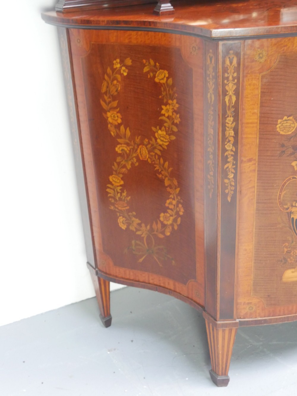 An antique mahogany display cabinet featuring marquetry urn inlays on lower doors, curved bowfront glass doors, glass shelves, and a swan-neck pediment. View VII