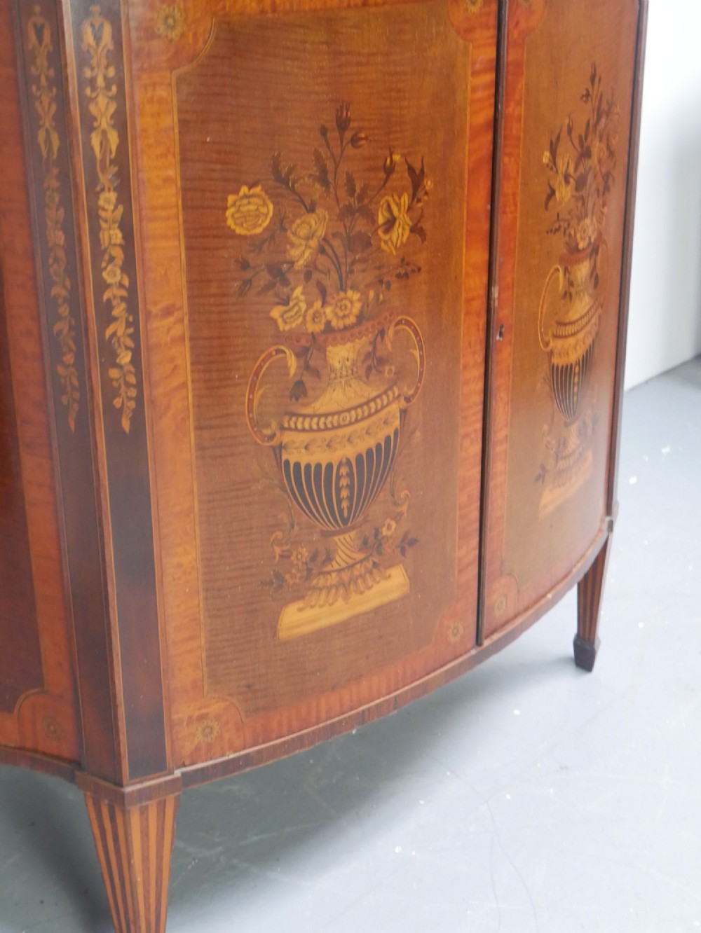 An antique mahogany display cabinet featuring marquetry urn inlays on lower doors, curved bowfront glass doors, glass shelves, and a swan-neck pediment. View VIII