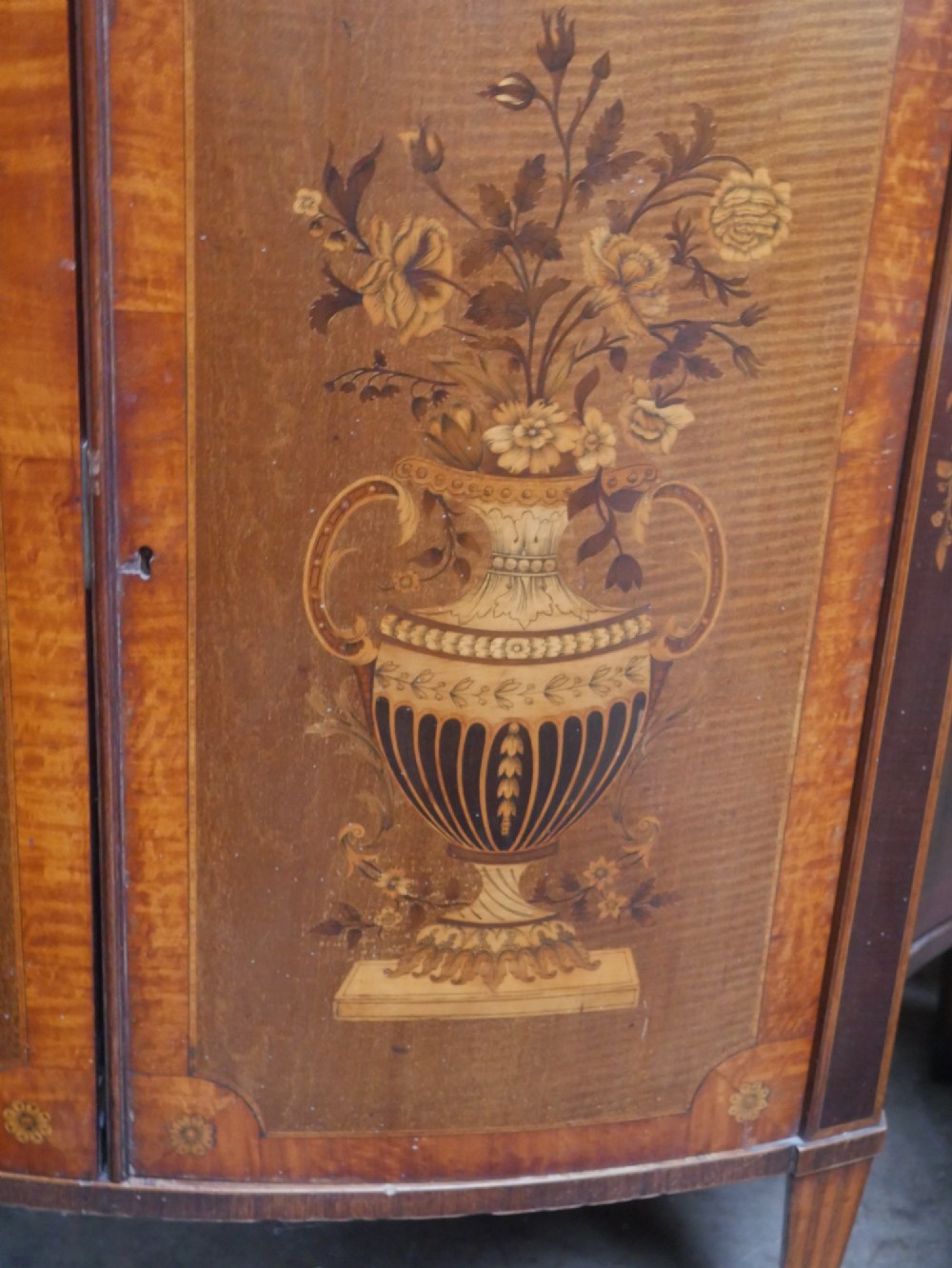 An antique mahogany display cabinet featuring marquetry urn inlays on lower doors, curved bowfront glass doors, glass shelves, and a swan-neck pediment. View X
