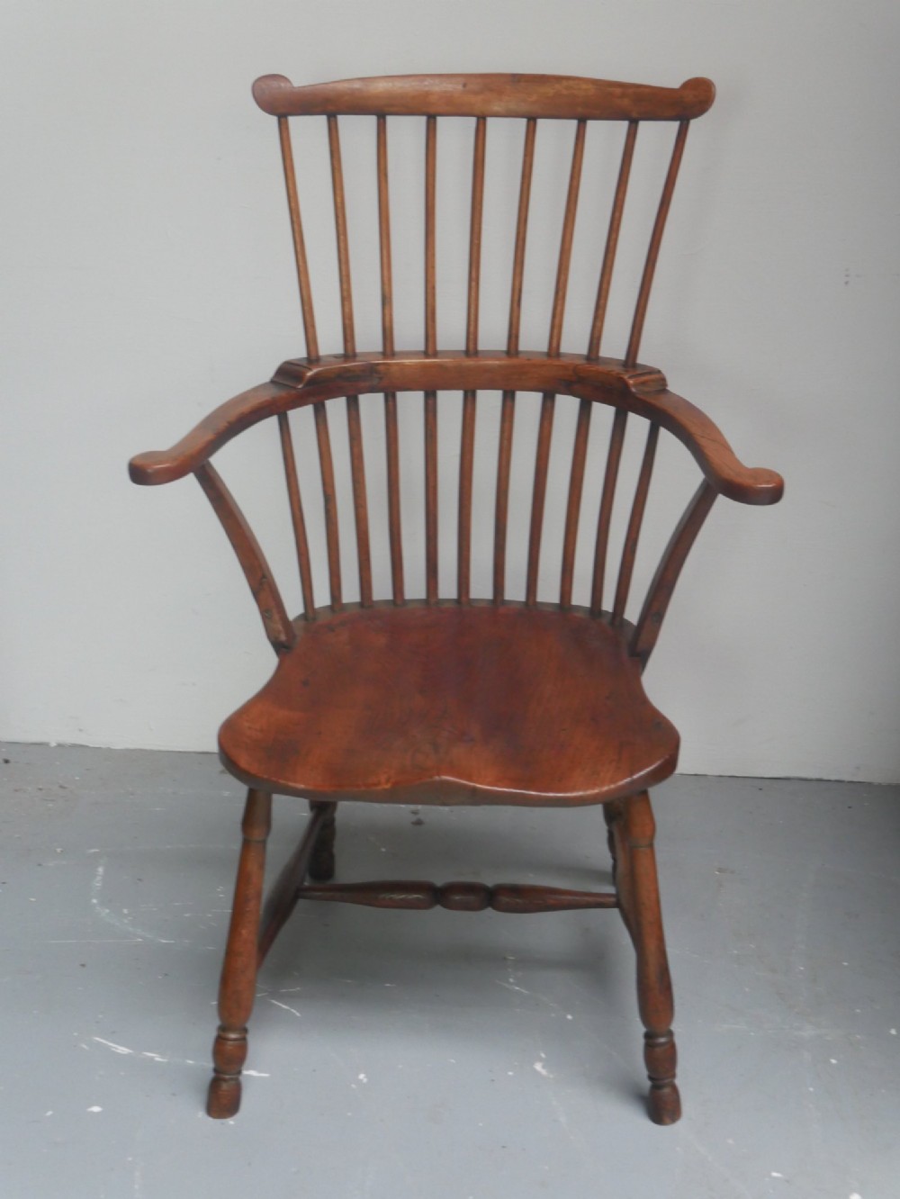 A mid-19th-century warm brown wooden Windsor armchair featuring a double-tier spindle back, carved saddle seat, curved armrests, and turned legs.