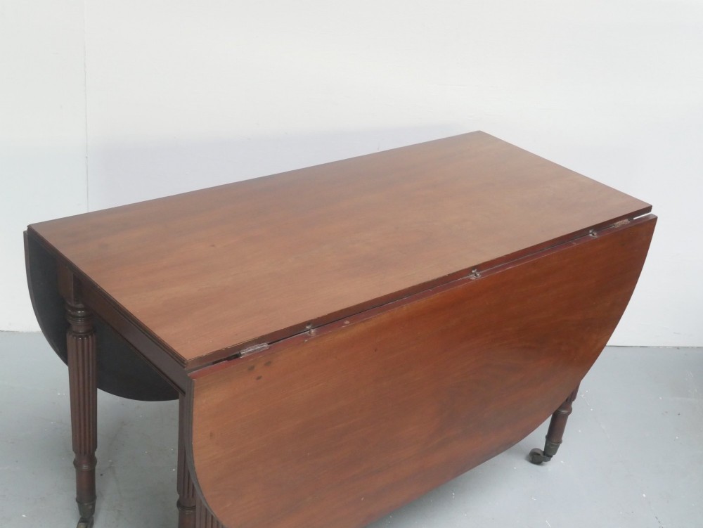 An antique mahogany drop-leaf dining table featuring reeded turned legs set on brass castors, displayed with both side leaves lowered. View II