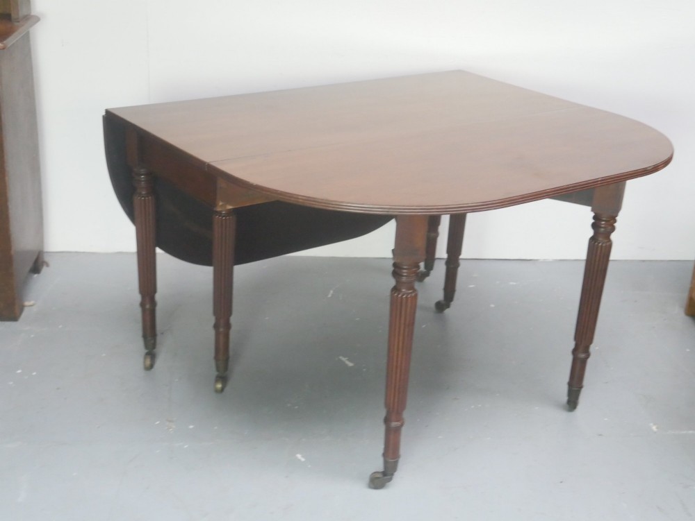 An antique mahogany drop-leaf dining table featuring reeded turned legs set on brass castors, displayed with both side leaves lowered. View VI