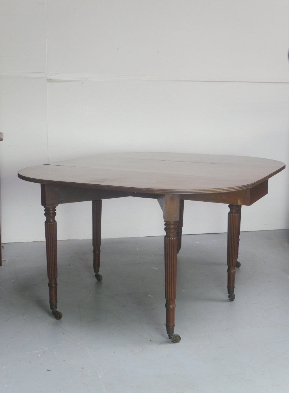 An antique mahogany drop-leaf dining table featuring reeded turned legs set on brass castors, displayed with both side leaves lowered. View VII