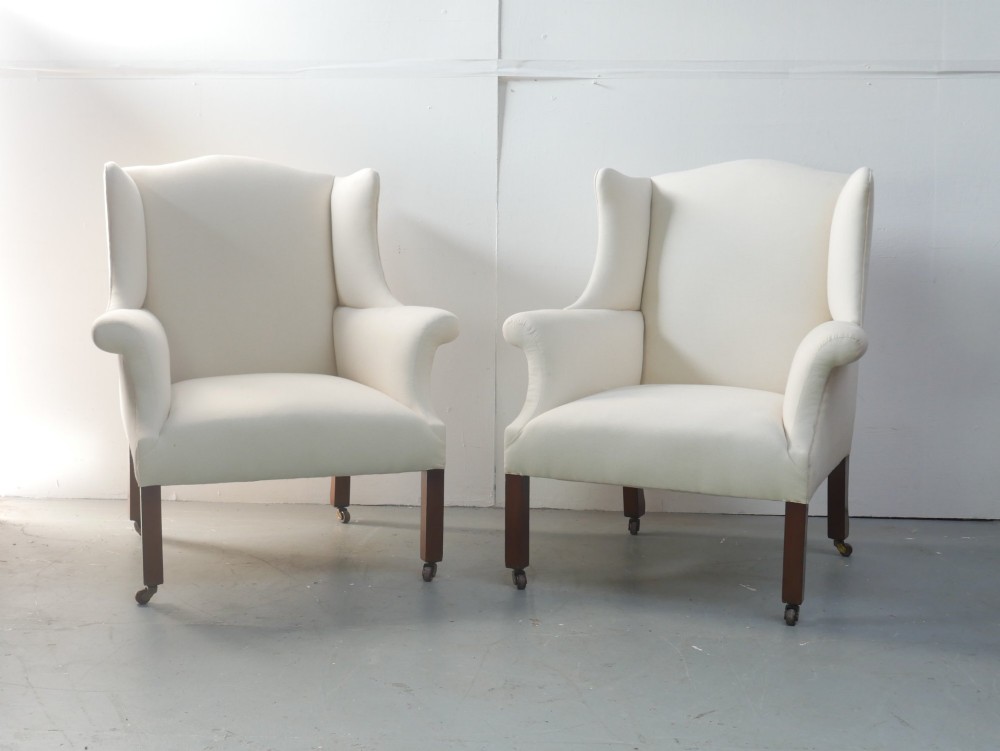 A pair of Edwardian wingback armchairs upholstered in cream fabric with square mahogany legs on brass castors.