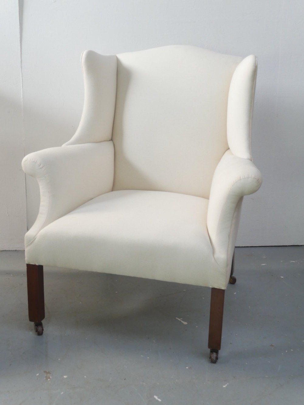 A pair of Edwardian wingback armchairs upholstered in cream fabric with square mahogany legs on brass castors. View II