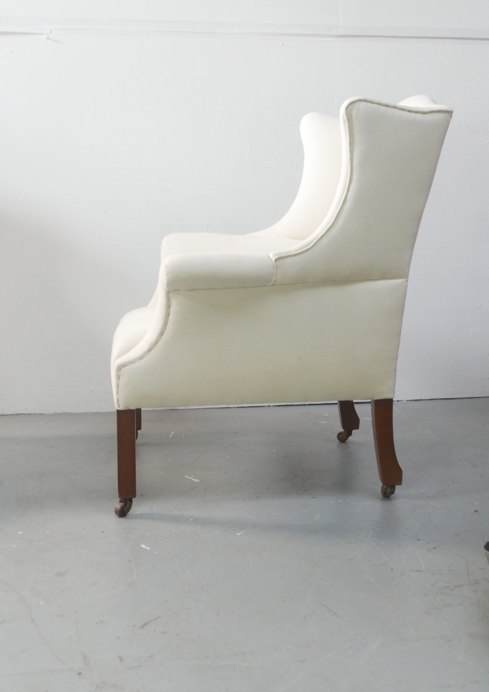 A pair of Edwardian wingback armchairs upholstered in cream fabric with square mahogany legs on brass castors. View III