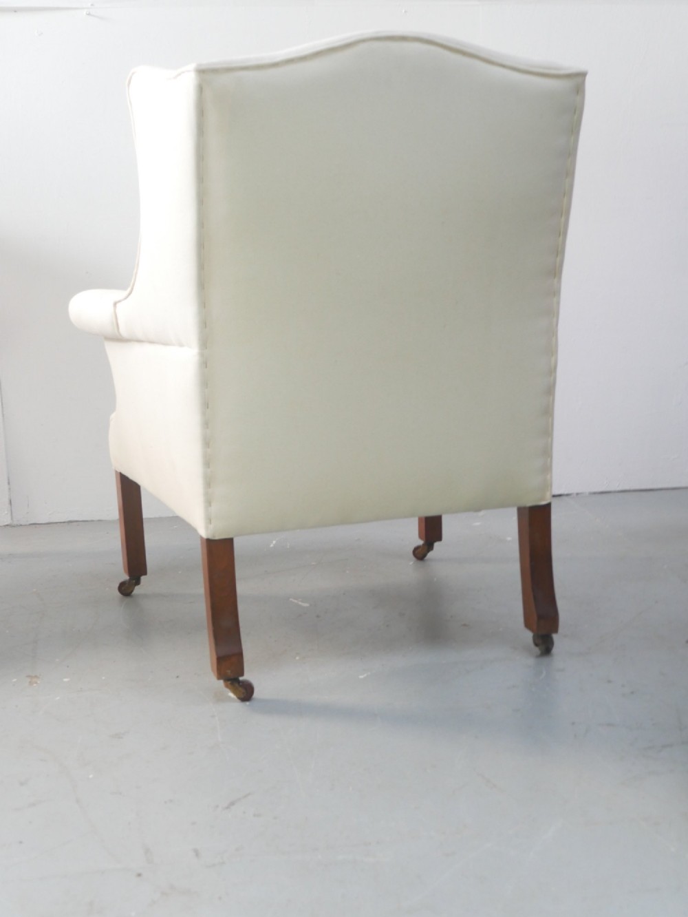 A pair of Edwardian wingback armchairs upholstered in cream fabric with square mahogany legs on brass castors. View IV