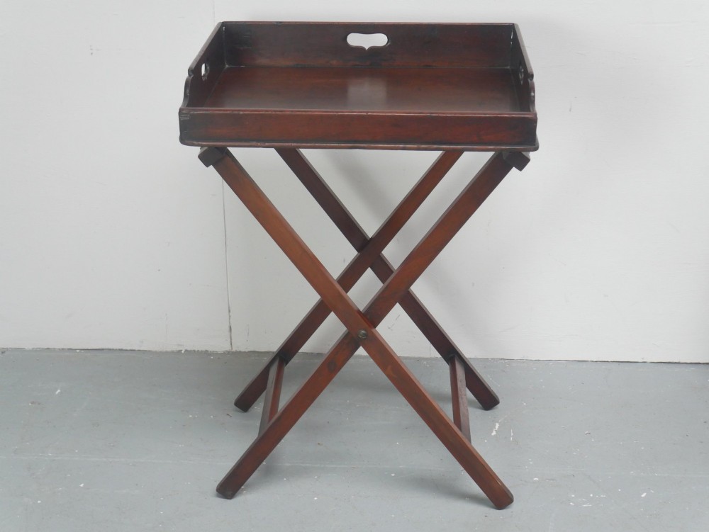A dark mahogany butlers tray with integrated hand cutout handles resting on a matching folding x-frame stand.